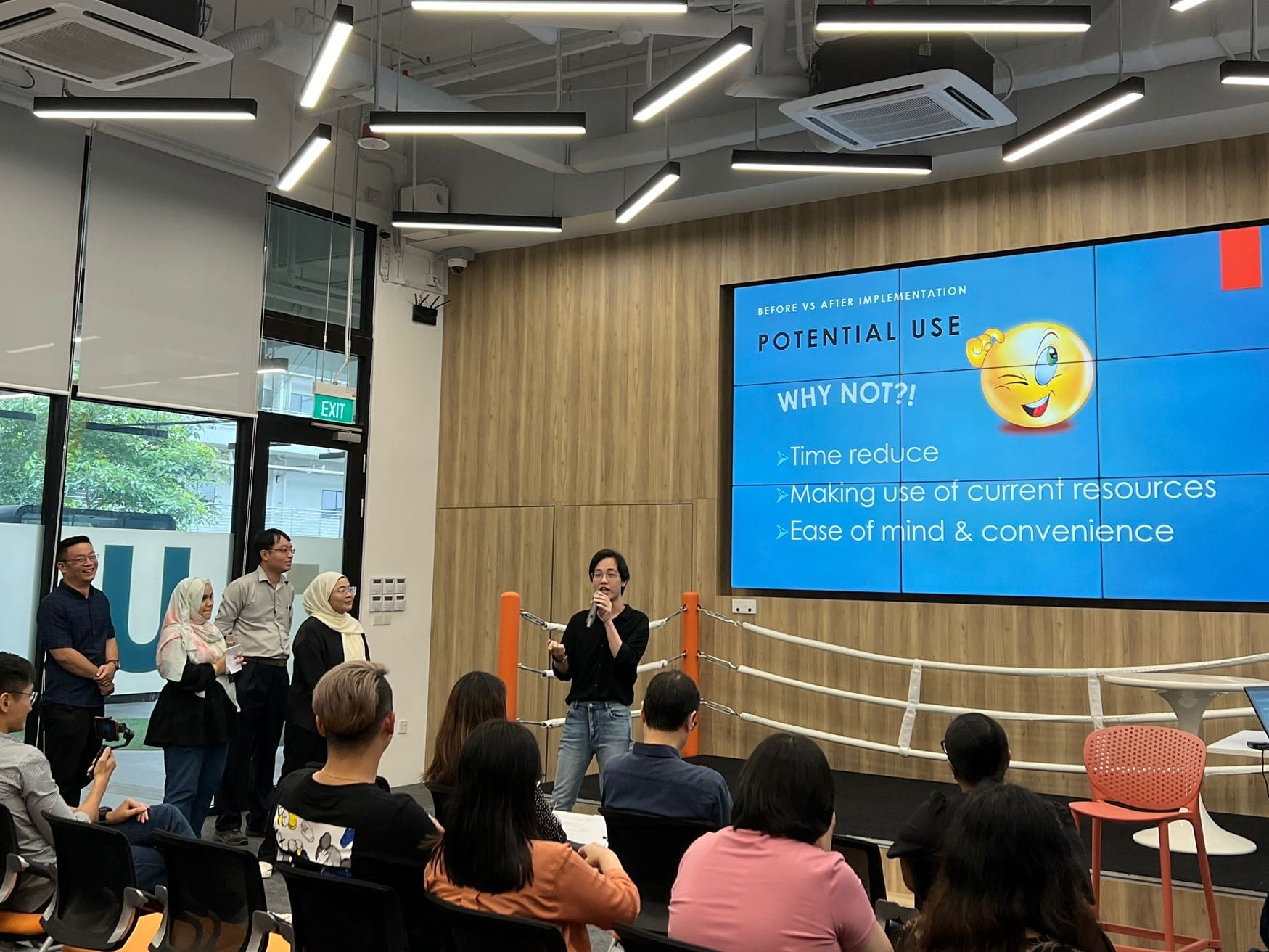 A presenter speaks before a group in a modern room, with a blue slide reading "Potential Use: Why Not?!" on a large screen.